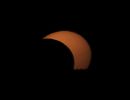 Ganz kurz vor Sonnenuntergang ist die jetzt intensiv orange schimmernde Sonne noch deutlich durch den Mond verdeckt und ein kleiner Teil ist schon unter den baumbestandenen Horizont abgetaucht. Partielle Sonnenfinsternis am 12. August 2026.